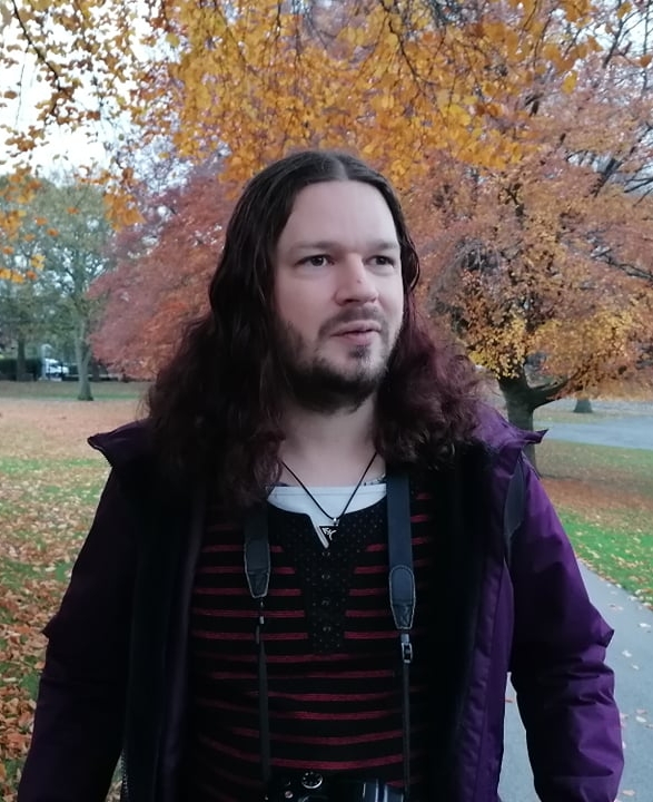 A photo of Steve, a white non-binary person in their late 30s, standing in a park with autumn trees behind them. Steve has long hair, dyed a purplish-red colour, and a short beard. They are wearing a purple coat with a red-and-black striped jumper underneath. Around their neck is a camera strap and a pendant with the transgender symbol on a black triangle.